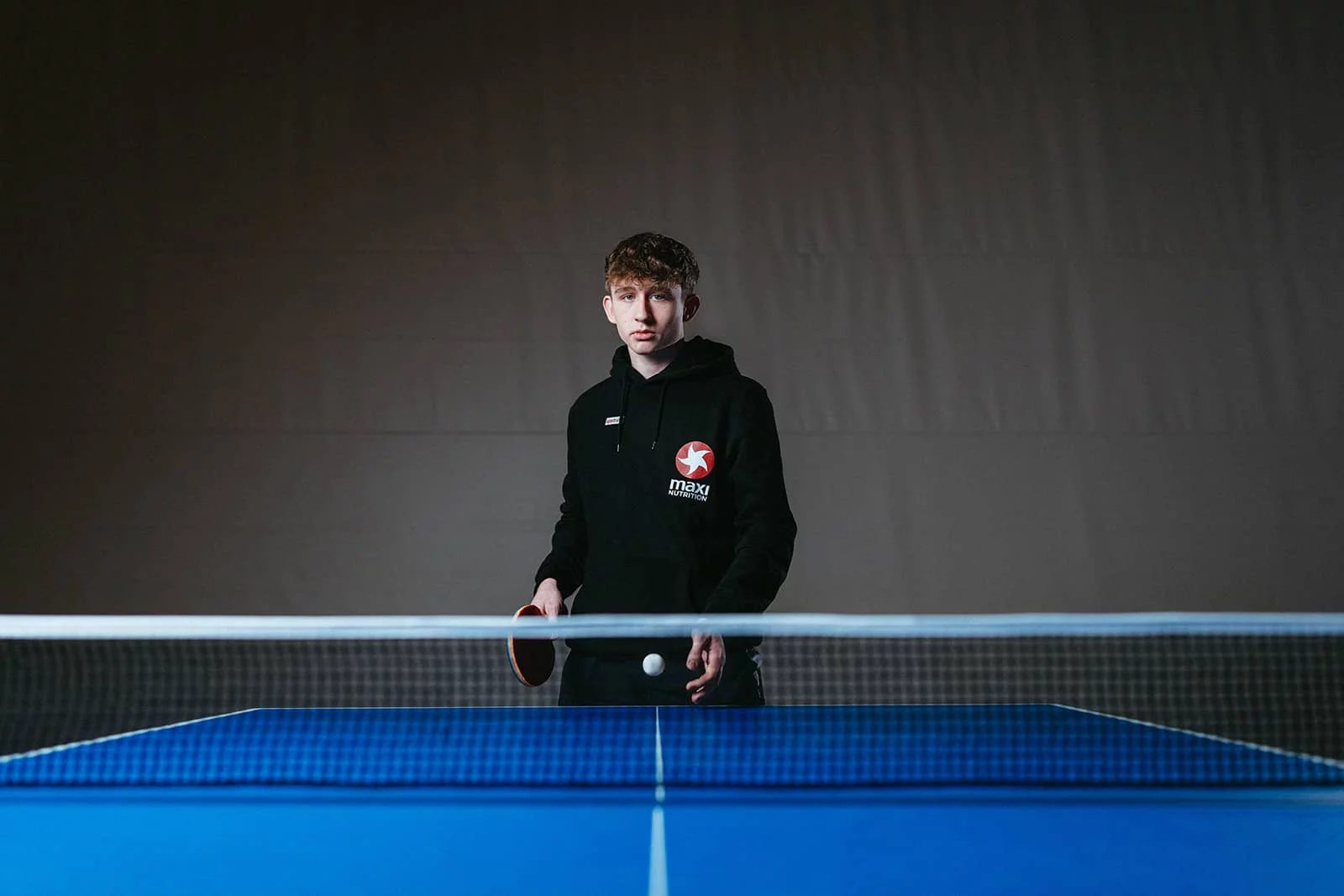 Julian Rzihauschek mit Schläger und Ball am Tischtennistisch im MaxiNutrition-Hoodie