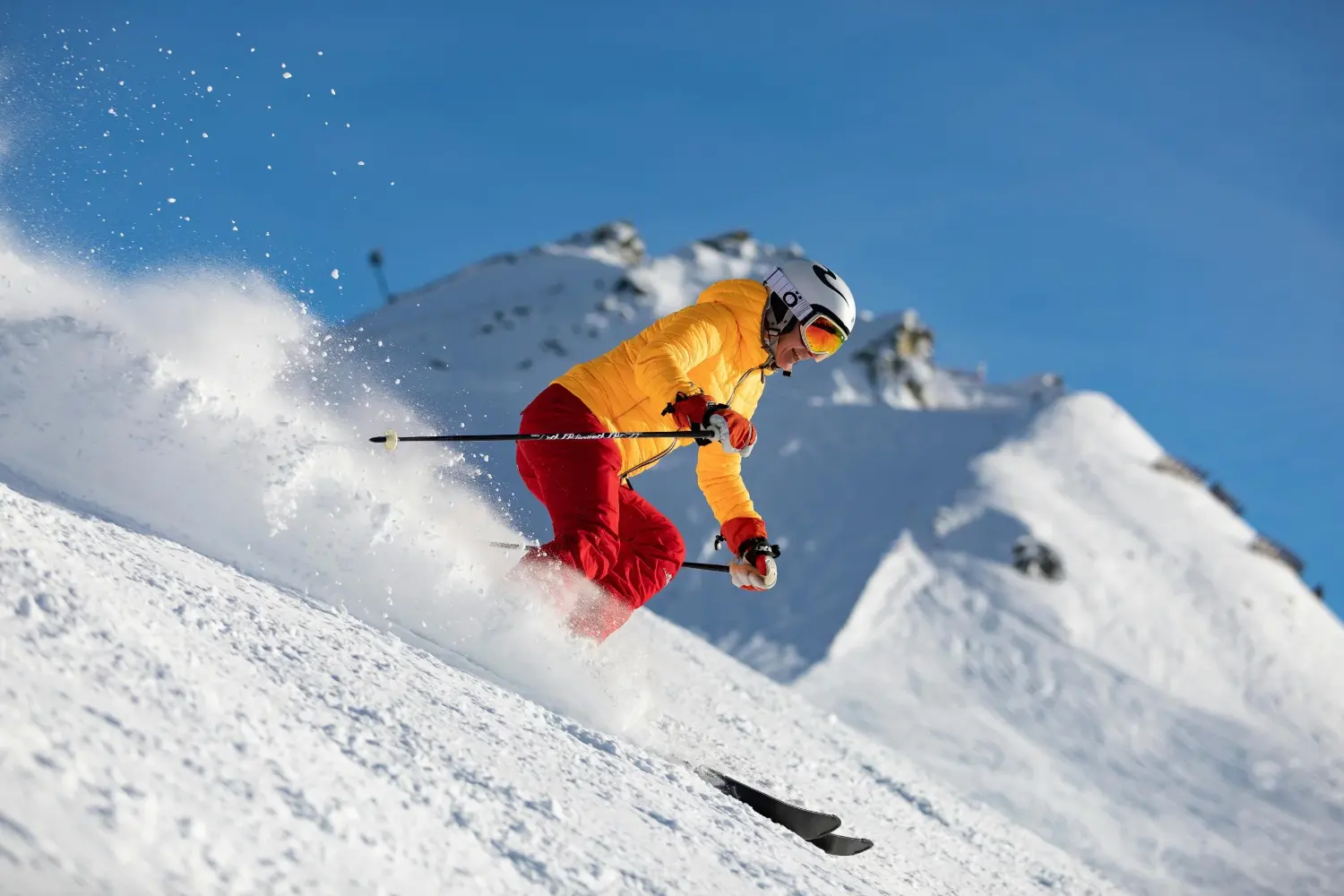 Skifahrer in gelber Jacke und roten Hosen carvet dynamisch durch Pulverschnee vor blauem Winterhimmel