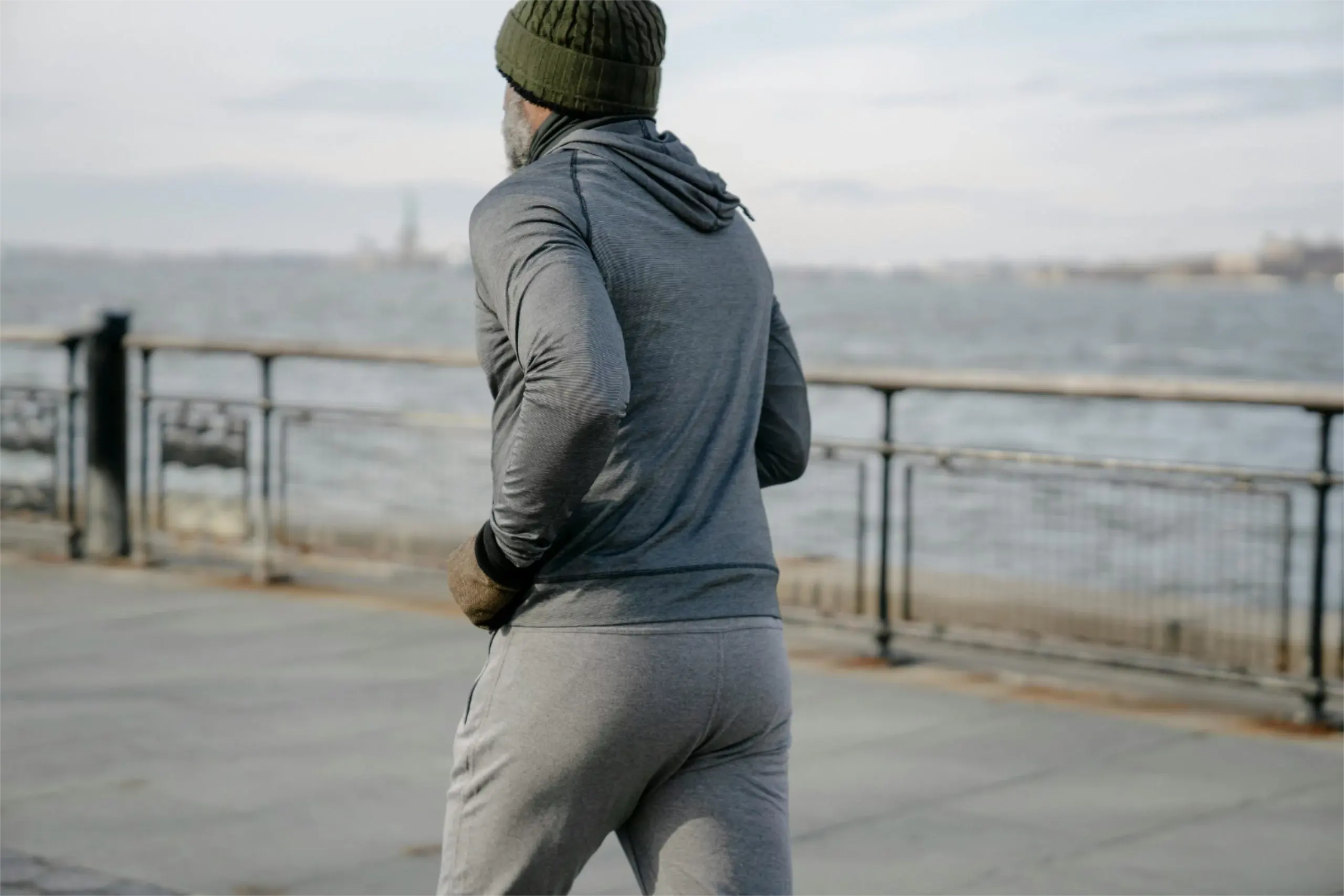 Mann in grauem Sportoutfit und Wollmütze joggt im Winter an einem Ufer entlang