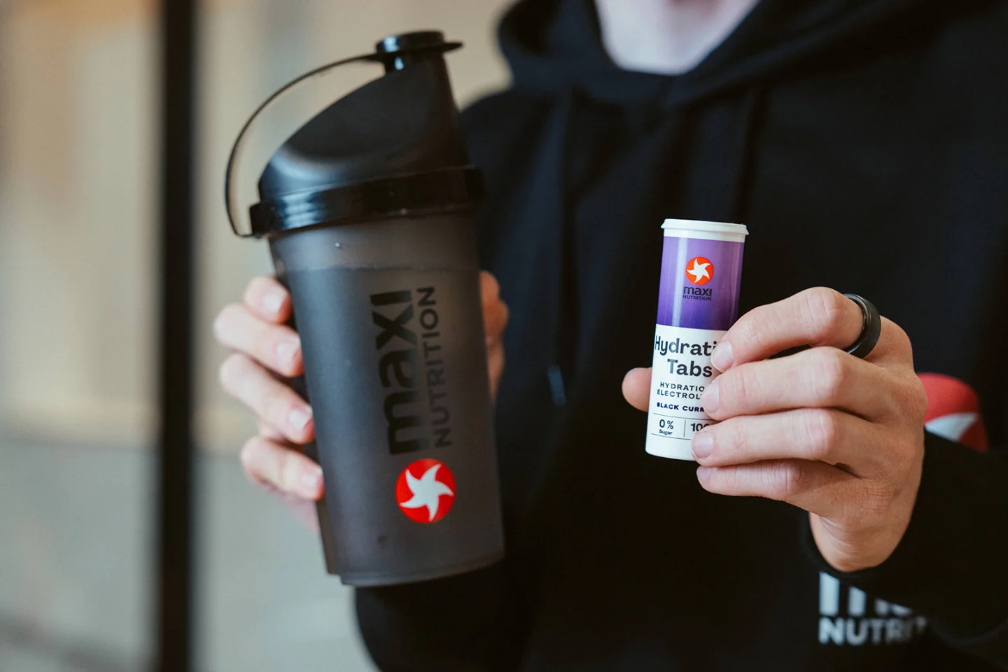 Sportler im MaxiNutrition-Hoodie hält schwarzen MaxiNutrition-Shaker und Hydration Tabs Black Currant in den Händen
