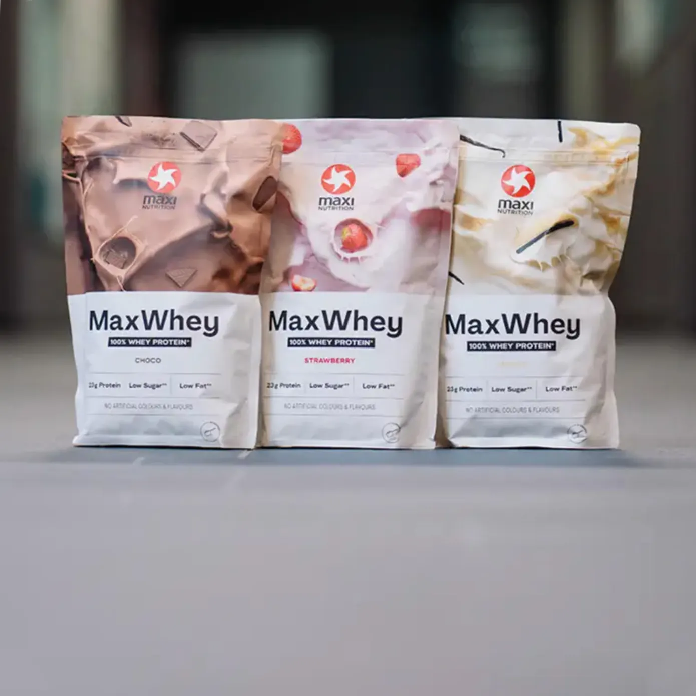 Drei Packungen MaxiNutrition MaxWhey 100% Whey Protein in den Sorten Choco, Strawberry und Vanilla nebeneinander
