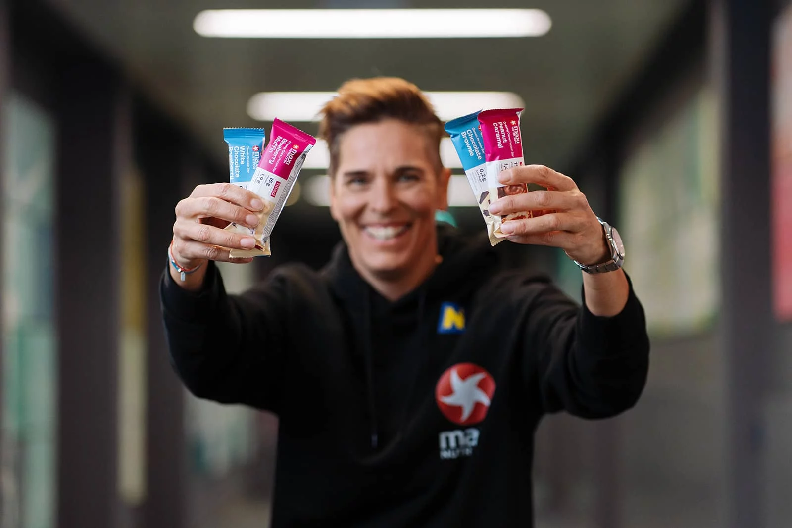 Michaela Dorfmeister im schwarzen MaxiNutrition Hoodie hält lachend vier Proteinriegel in die Kamera
