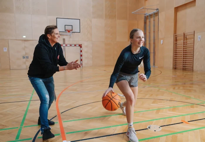 Mentorin feuert lächelnd ein junges Basketballtalent beim Dribbling in einer Sporthalle an
