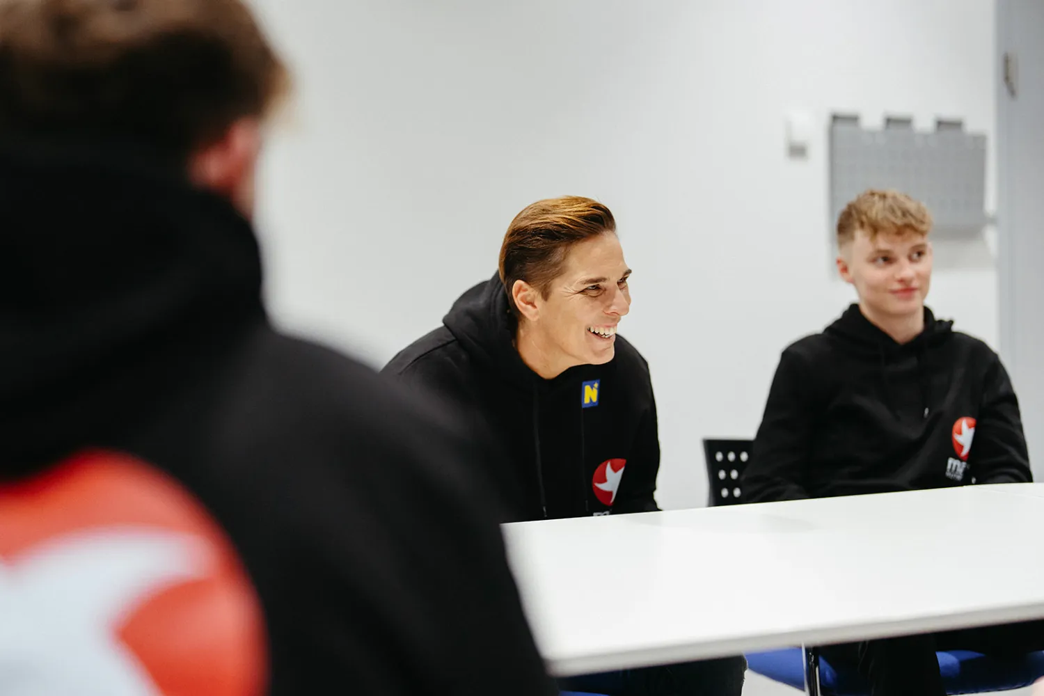 Mentorin lacht beim Mentoring-Gespräch mit jungen Sporttalenten in schwarzen MaxiNutrition-Hoodies am Tisch