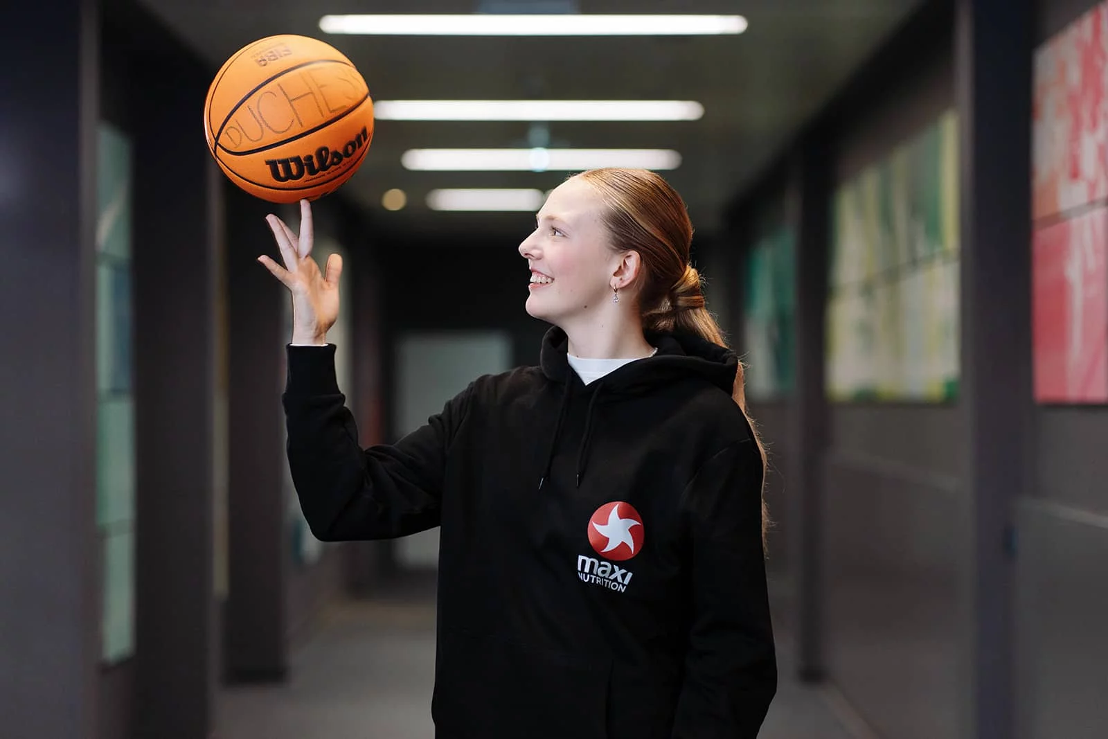 Sina Höllerl im MaxiNutrition-Hoodie balanciert lächelnd einen Wilson-Basketball auf dem Finger