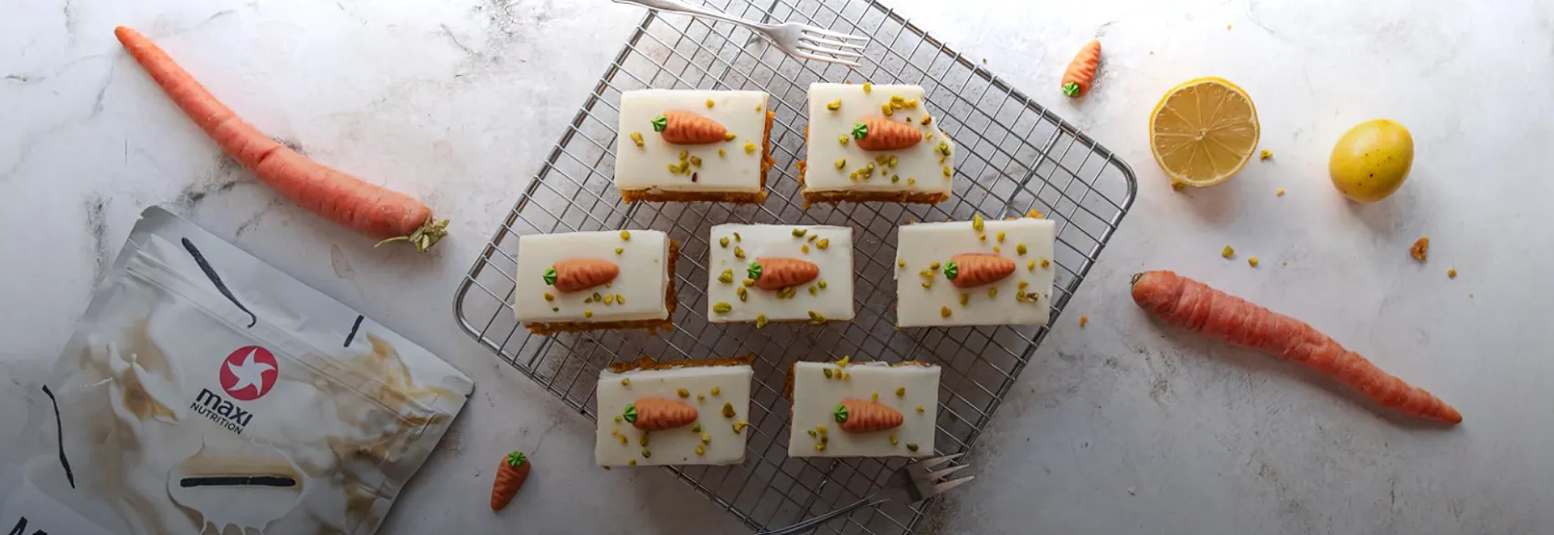 Sechs Stücke Karottenkuchen mit weißem Frischkäse-Frosting und Karottendeko auf einem Kuchengitter, daneben MaxiNutrition Proteinpulver
