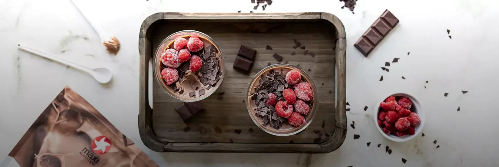 Zwei Gläser Protein Mousse au Chocolat mit gefrorenen Himbeeren und Schokoladenstücken auf einem Holztablett