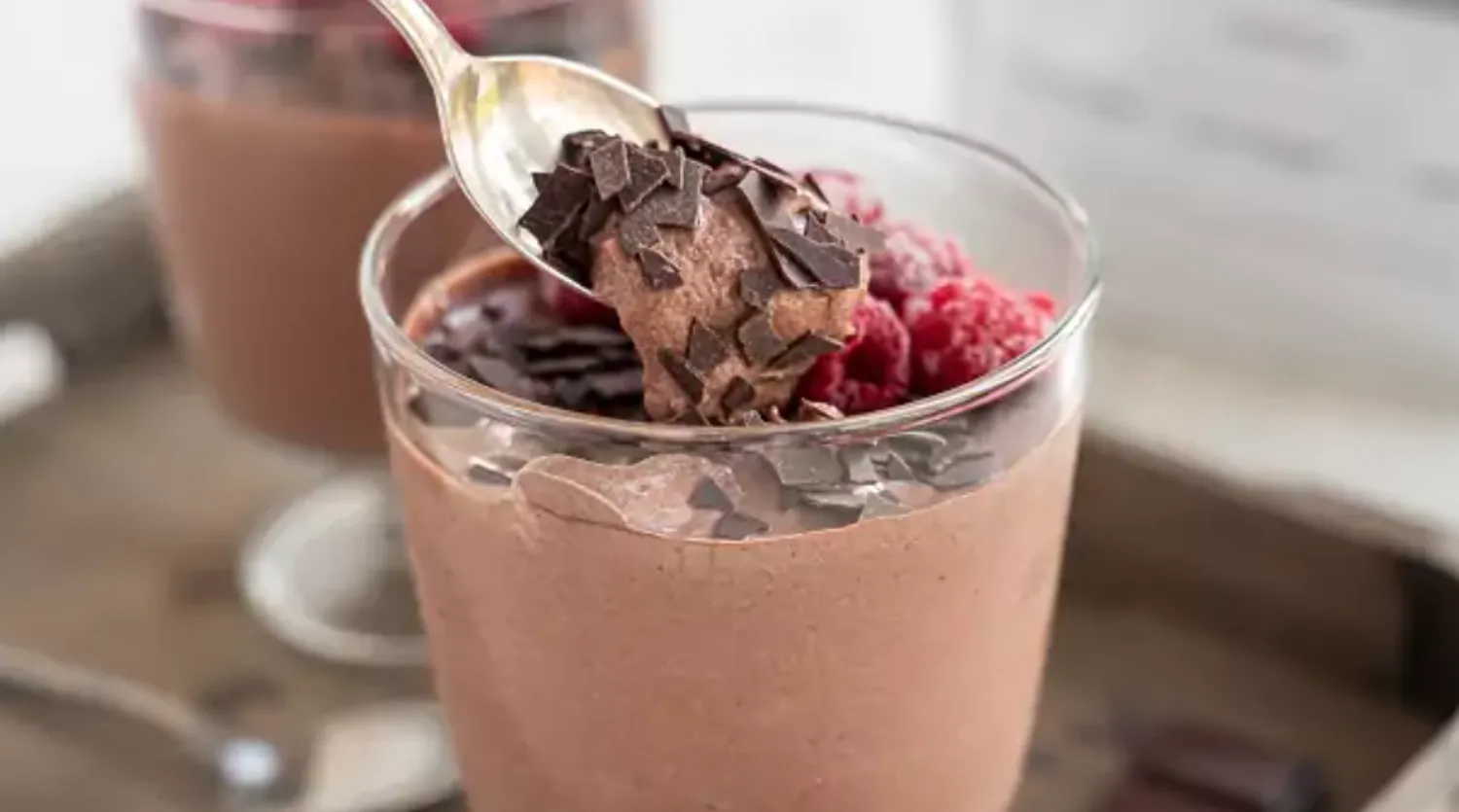 Nahaufnahme eines Löffels mit cremiger Protein Mousse au Chocolat mit Schokoladenstücken und Himbeeren im Glas