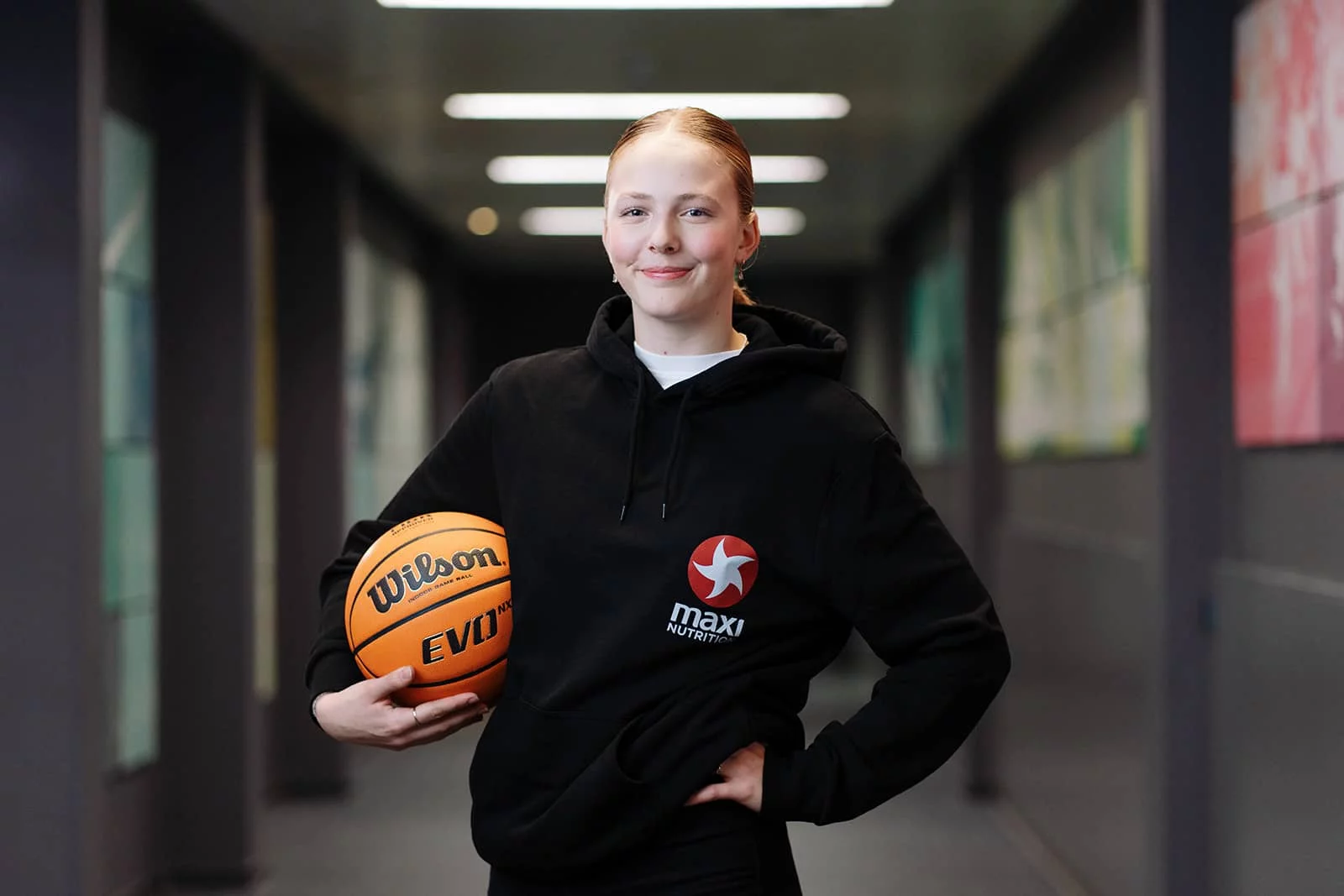 Sina Höllerl steht in einem Schulgang mit einem Basketball unter dem rechten Arm.